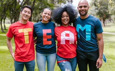 A diverse group of adults bonding and smiling in a park wearing TEAM shirts.
